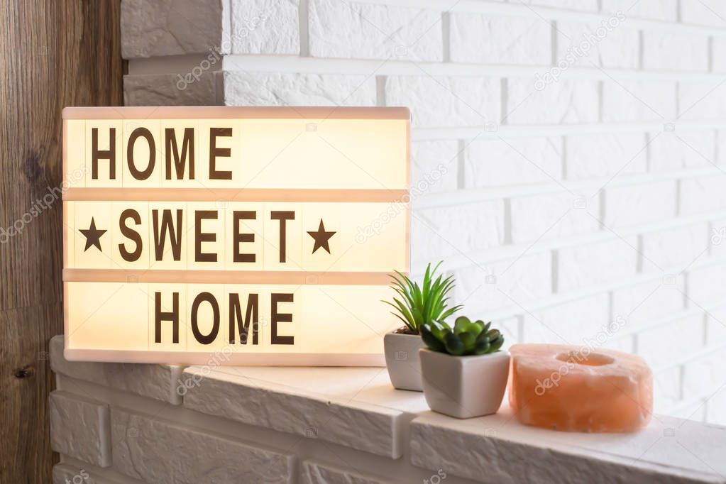 Home sweet home - it is written on a decorative frame with illumination in the interior between a white brick wall and a brown wooden one next to succulents and a pink candlestick. Details and decor of the interior
