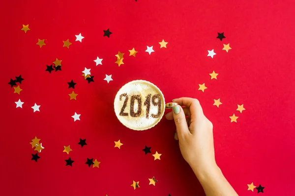 Yeni yıl 2019 bir fincan sütlü kahve üzerinde bulunmakta. 1 Ocak yeni yıl kavramı. Parlak yıldız ile kırmızı zemin üzerine. Üstten görünüm, düz yatıyordu