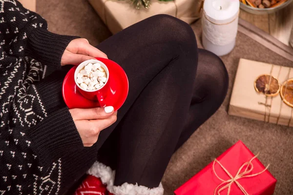 A small red cup with hot chocolate and marshmallows in the hands of a woman in red warm socks sitting among packed Christmas gifts Top view flat lay