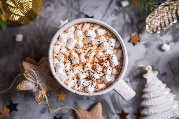 A large cup of cocoa with marshmallow sprinkled with cocoa powder stands on a gray table among Christmas decorations, fir branches, ginger cookies and shiny stars. Top view