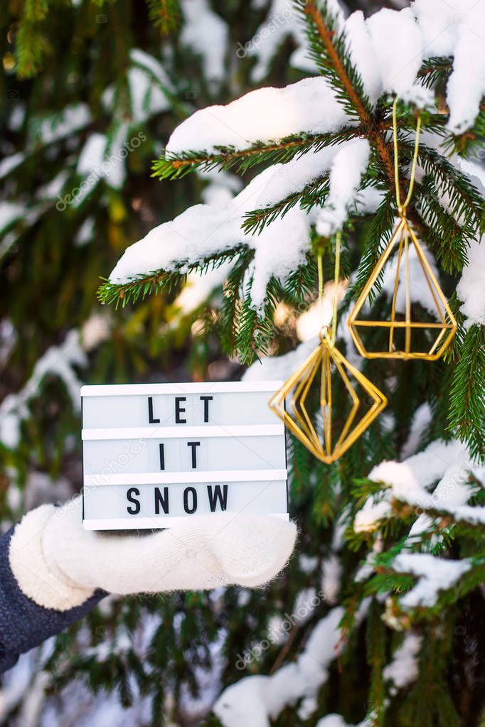 Let it snow it is written on a decorative lamp in the hand of a woman in a white mitten against the background of green fir branches in the snow
