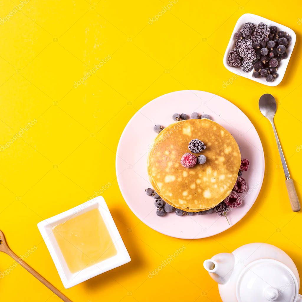 A stack of pancakes on a pink plate with berries next to a plate of honey and jam with wooden spoons on a bright yellow background. Top view, copy space