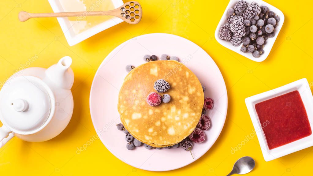 A stack of pancakes on a pink plate with berries of blackberries, raspberries and blackberries next to honey, jam and a kettle of tea on a bright yellow background