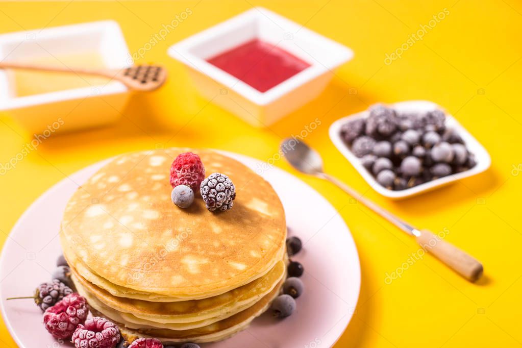 A stack of pancakes on a pink plate with berries next to honey and strawberry jam on a bright yellow background