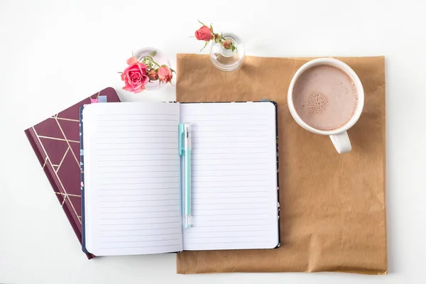 Açık bir defter bir vazo gül ve bir fincan kahve yanında beyaz bir masaüstünde boş çarşafları ile. Üstten görünüm, düz yatıyordu