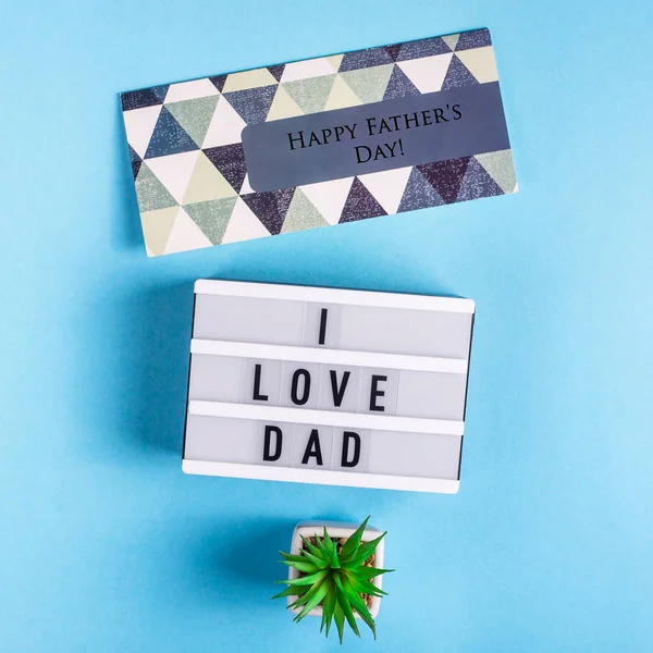 Father\'s day layout with a postcard and the inscription I love dad on a decorative lamp next to a pot of succulents on a blue background. Top view, flat lay