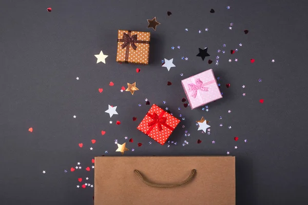 Kraft paper bag on bright dark background. With gift boxes and confetti. Black friday christmas gift preparation concept.