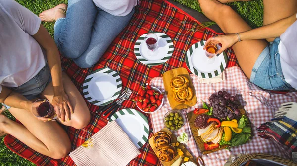 Three girls have picnic with chopped meat and cheese, fruits, grapes, strawberries and buns with glasses of wine on a red checkered.picnic sheet