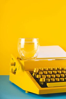 Sarı parlak daktilo, sarı ve mavi arka planda, bir bardak suyun yanında. Yazma, blog yazma, yeni fikirler ve yaratıcılık için sembol