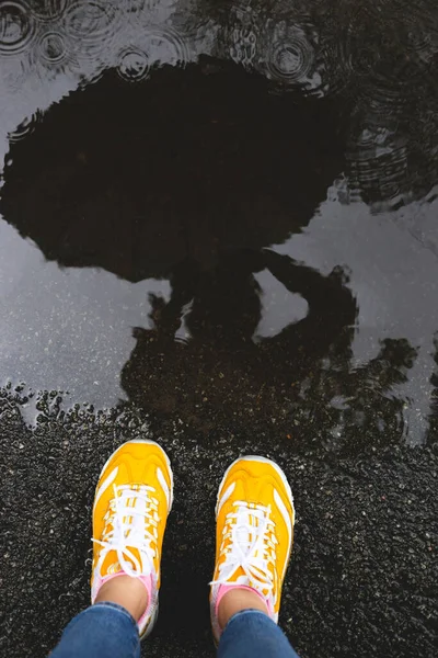 Woman with umbrella is reflected in a puddle. Bright yellow sneakers and raindrops on the asphalt