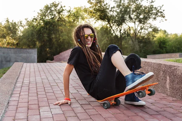 dreadlocks güneş gözlüğü ile moda kız kaykay ve gülüyor düştü. uzun bir tahta ile bir parkta olumlu kız hippi. hipster tahta üzerinde paten öğrenmek.