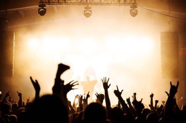 Sahnedeki müzik grubundan bir vokalistin silueti ve konserdeki keçi kalabalığı. Turuncu ışıklar altında festival posteri.