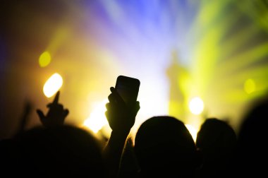 Bir hayranın elinde akıllı telefon, sevilen bir sanatçının konserinde hatıra için video ve fotoğraf çekiyor. Işıklı bir müzik festivalinin atmosferi. Pankartı göster