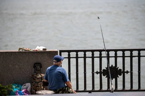 保護マスクとキャップの高齢漁師が座って 市内の川の堤防で魚をキャッチします 戦利品と装備袋 — ストック写真