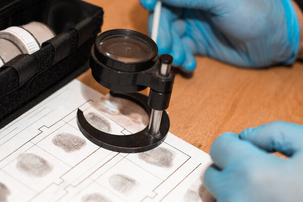 investigator or detective in rubber disposable gloves examines fingerprints on paper through a magnifying glass. detective agency office. crime investigation