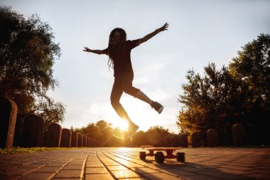 Parkta batan güneşin altında kaykay üzerinde zıplayan bir kızın silueti. Rastalı ve güneş gözlüklü hippi uzun tahta üzerinde numaralar öğreniyor..