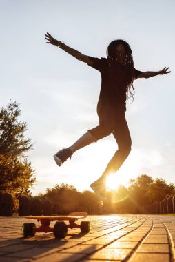 Parkta batan güneşin altında kaykay üzerinde zıplayan bir kızın silueti. Rastalı ve güneş gözlüklü hippi uzun tahta üzerinde numaralar öğreniyor..