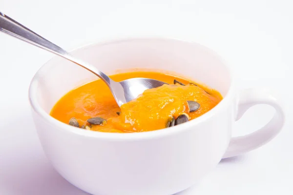 白い背景の上のスプーンで食べられているかぼちゃの種で飾られたカボチャのスープ — ストック写真