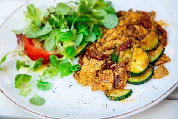 Vegan breakfast prepared with scrambled tofu and eggs, cooked with dried tomatoes and zucchini served with fresh lettuce and cucumber in white ceramic vintage plate