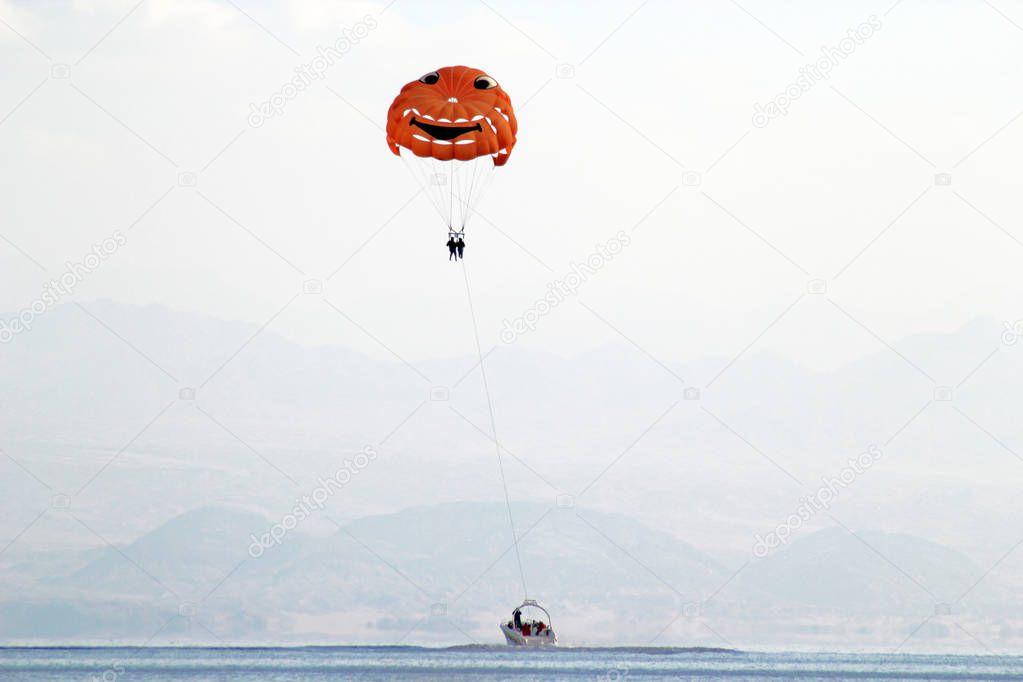 orange paraglider with a cheerful pattern hanging over a boat against a background of mountains in the haze
