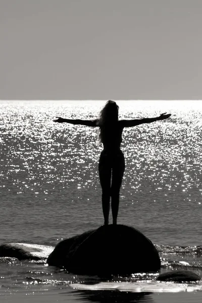 Foto di una ragazza silhouette in piedi su una pietra con le braccia divaricate. Vicino al mare. Contro il bagliore splendente del sole sulla superficie del mare . — Foto Stock