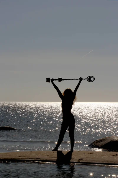 一个女孩在沙滩上的剪影，她手拿金属探测器举过头顶。与海面上闪闪发光的太阳眩光。计数器拍摄. — 图库照片