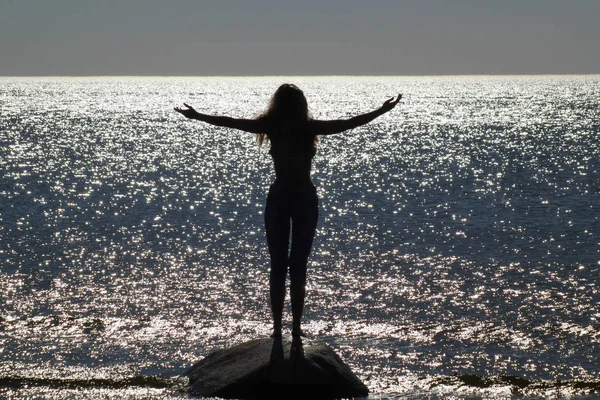 Kolları dağılmış bir taşın üzerinde duran siluetli bir kızın fotoğrafı. Denizin yanında. Deniz yüzeyinde güneşin parıldayan parıltısına karşı. — Stok fotoğraf