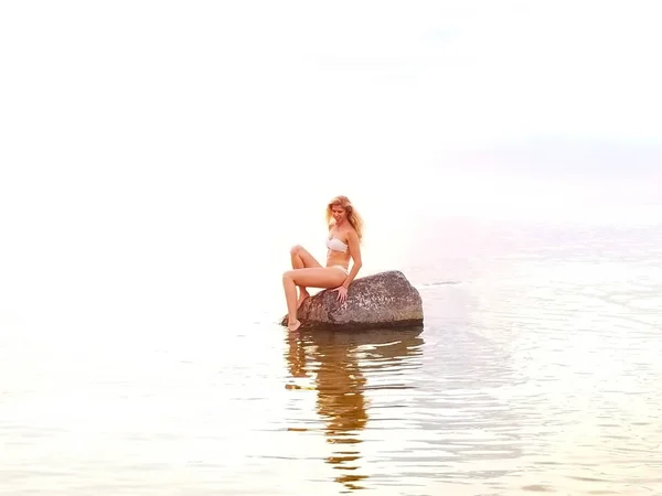 Hermosa rubia en un bikini blanco se sienta en una piedra de pie en el agua durante la puesta del sol — Foto de Stock