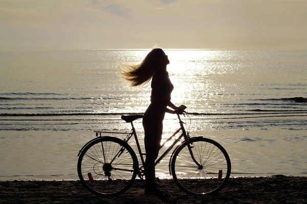 Silhouette of a girl with long hair on a bicycle riding on the sand by the Baltic sea. Shooting against the sun. — Stock Photo, Image