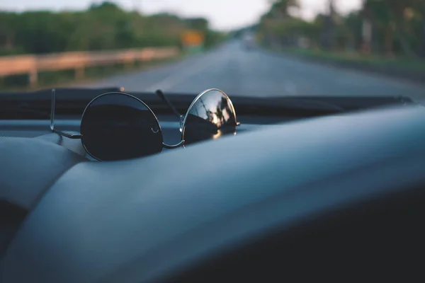 Sunglasses on car panel with view of road background while traveling on summer vacation with sunset reflection on glasses