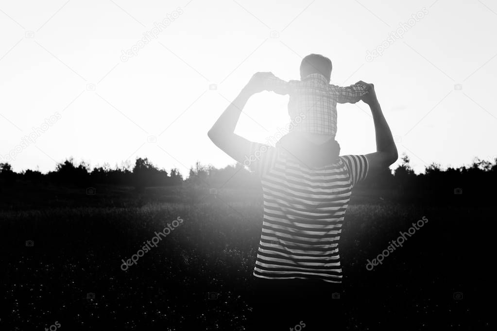 Back view of happy little boy stretching out hands while his father carrying him on shoulders. Silhouette portrait on sunset background