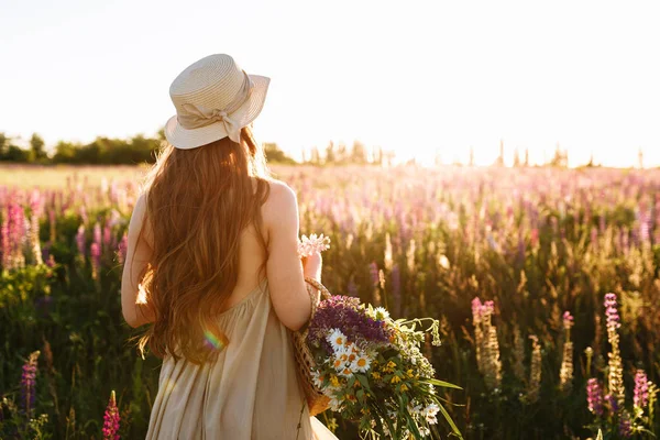 Giovane donna in cappello di paglia e vestito con bouquet di fiori di lupino, vista posteriore in campo tramonto — Foto Stock