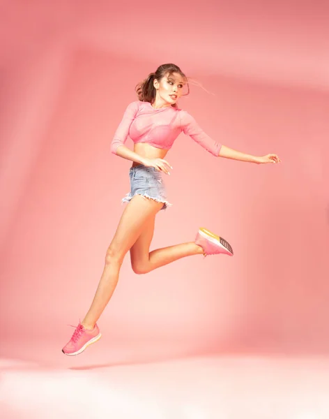 sporty woman with horse tail hairstyle in summer clothes jumping on pink background