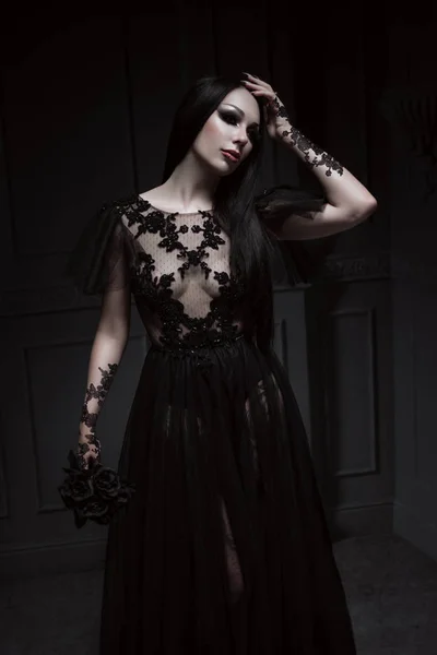 美丽的黑发女人与哥特式化妆穿着时尚的黑色连衣裙和在灰色背景上摆姿势 — 图库照片