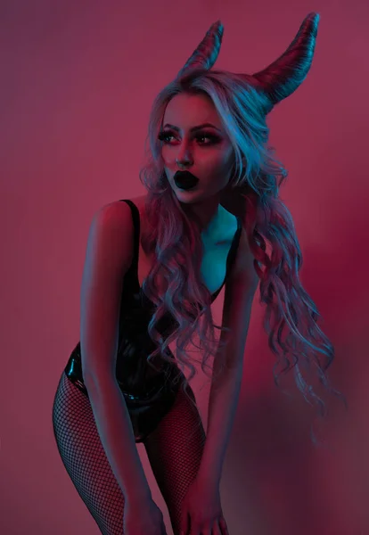 ダークピンクの背景にポーズ黒いラテックスボディを身に着けている悪魔の角を持つ魅力的なブロンドの女性 — ストック写真