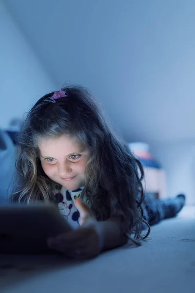 在床上用数码平板电脑 晚上看电影或玩游戏 微笑着快乐可爱的女孩 一头长长的卷曲的金发 — 图库照片