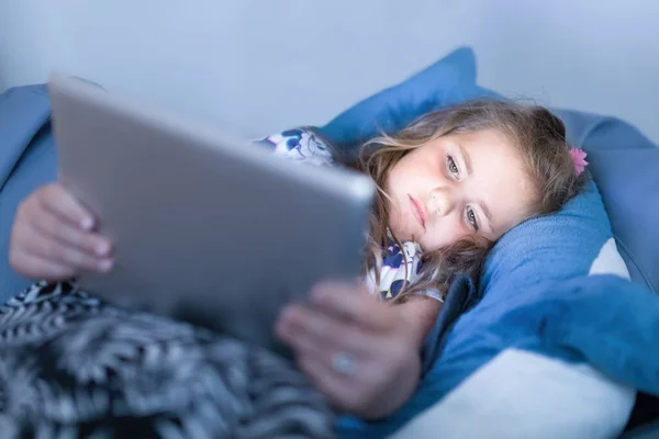 以在床上使用数码平板电脑 晚上看电影或玩游戏的可爱小女孩为重点 — 图库照片