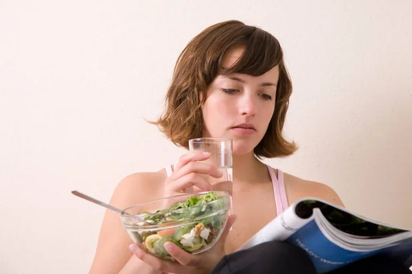 かなりブルネット座っているソファの上に雑誌を読んでいる間にサラダを食べ 彼女の手に水のガラスを持っている — ストック写真