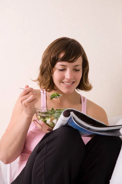 漂亮的黑发女人坐在沙发上放松地看杂志 吃沙拉 — 图库照片
