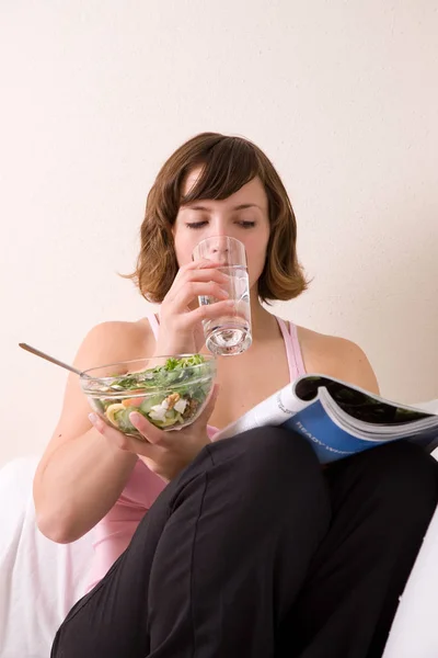 穿着运动服坐在沙发上的漂亮的黑头发姑娘 吃着沙拉 喝着一杯水 — 图库照片