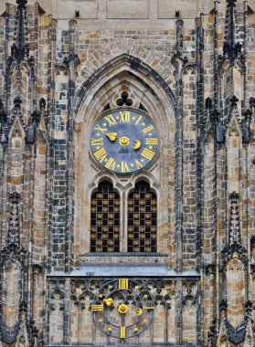 PRAGUE, CZECH REPUBLIC - May 2, 2018: Fragment of the wall of Saint Vitus cathedral with a window protected by a gilded curved lattice, and a tower clock.