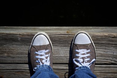 Looking down at sneaker clad feet at edge of pier, water is dark like looking into the abyss, light is bright sunshine.