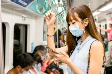 Genç Asyalı kadın yolcu ameliyat maskesi takıyor ve metroda cep telefonuyla müzik dinliyor.