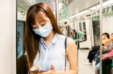 Genç Asyalı kadın yolcu ameliyat maskesi takıyor ve metroda cep telefonu kullanıyor.