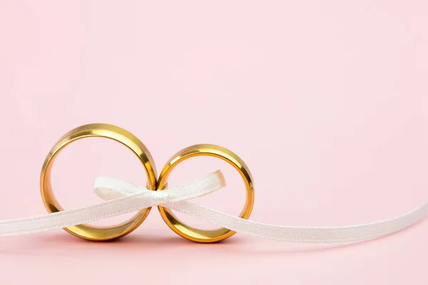 优雅的婚礼或订婚背景 一对金色的结婚戒指和柔和粉色背景的柔和的白色蝴蝶结带 复制文字空间 — 图库照片