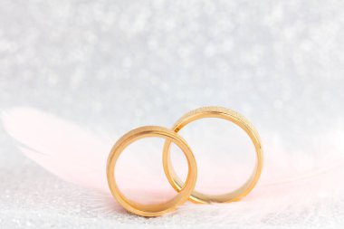 İki Altın Nikah Yüzüğü ve Hafif Melek Tüyü, yumuşak, ışıl ışıl arka planda.