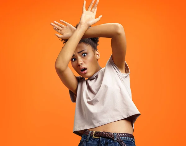 Scared girl outstretched her palms, hiding her face. Photo of african american girl wears casual outfit on orange background. Emotions and feelings concept.