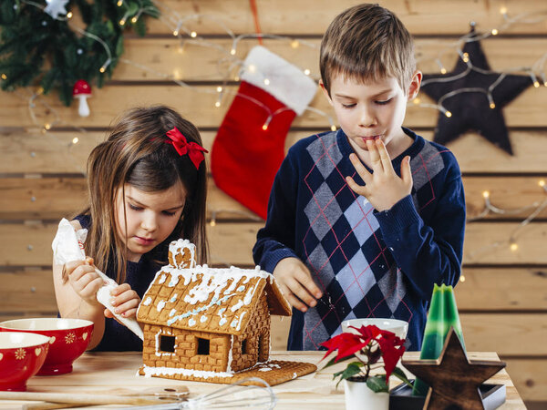 Photo of a young brother and sister decorating a gingerbread house at home just before Christmas.