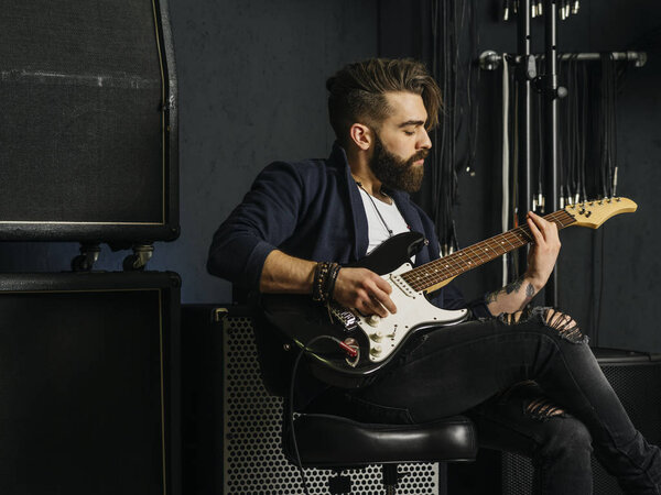 Photo of a man with beard sitting and playing his electric guitar in a recording studio.