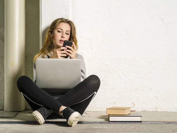 Studentka sedí se svým notebookem a dívá se na svůj smartphone — Stock fotografie